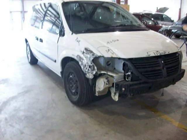 A white Dodge Caravan on the C & C Collision Center shop floor with front-end collision damage, the bumper and headlamp removed