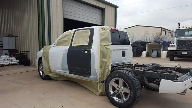 A white Ram crew cab masked in paper with the bed removed, standing outside the C & C Collision Center bay door
